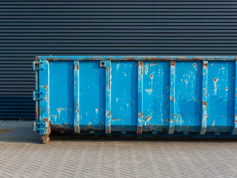 A blue steel roll-off dumpster standing on brick paving in front of a dark metal wall