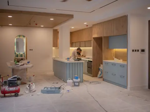 A kitchen being renovated: new wood and blue cabinets, a covered floor, a compressor and tools, and a worker at the counter